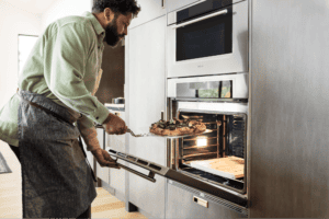 A man wearing an apron uses a pizza peel to place a cooked pizza into a stainless steel Wolf oven in a modern kitchen equipped with luxury kitchen appliances.