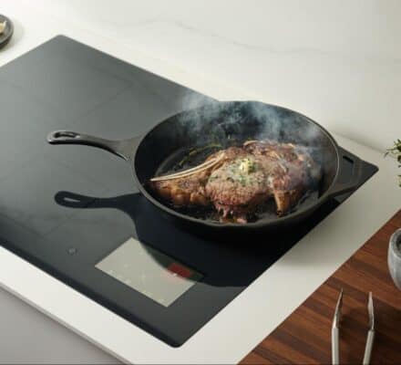 A cast iron skillet with a steak sizzling on an induction cooktop, steam rising as fresh herbs, salt, and tongs rest nearby—illustrating how to use induction cooking for perfect results on a wooden counter.