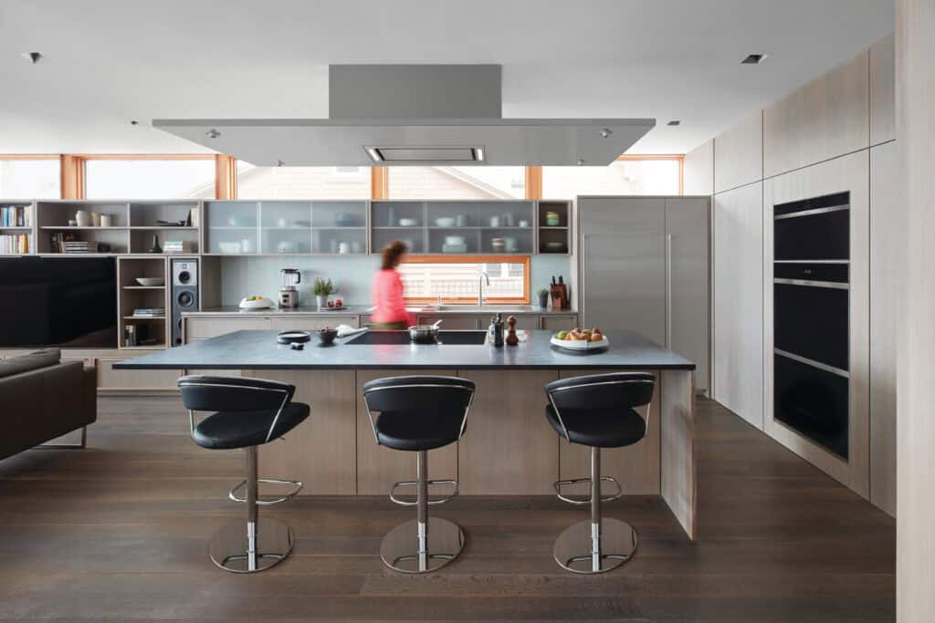 Modern kitchen with a large island, three bar stools, built-in appliances, and a person in a pink top blurred in motion. Shelves and cabinets line the walls—perfect inspiration for your next luxury kitchen renovation.