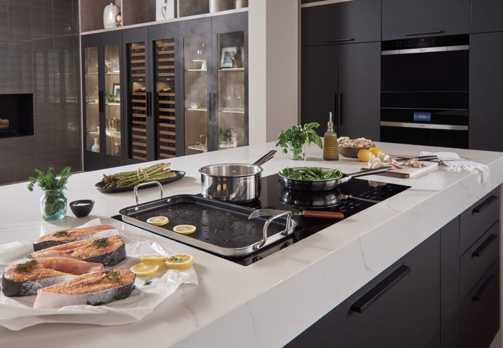 Modern kitchen with black cabinets and white countertops; Wolf kitchen appliances shine as salmon steaks, vegetables, herbs, and utensils are arranged on the counter beside a busy stovetop—just one of many reasons to invest in quality cooking spaces.