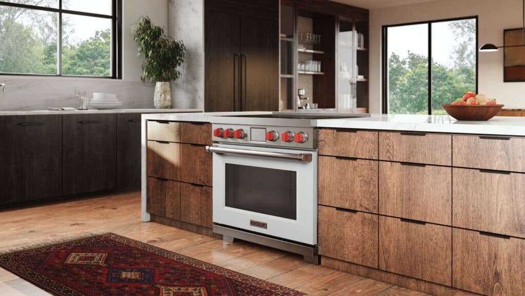 Modern kitchen with wood cabinets, a white oven with red knobs, wolf induction cooktop, marble countertop, large windows, and a patterned rug on the wooden floor.