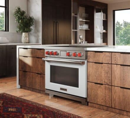 Modern kitchen with wood cabinets, a white oven with red knobs, wolf induction cooktop, marble countertop, large windows, and a patterned rug on the wooden floor.