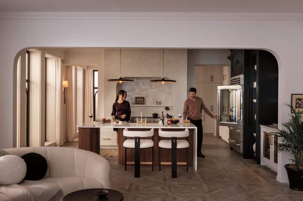A woman and a man prepare food in a luxury kitchen featuring an island, bar stools, pendant lights, and sleek stainless steel appliances.