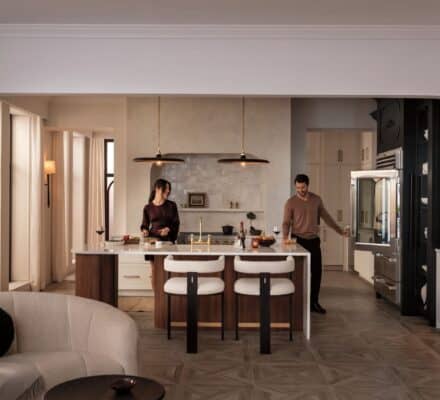 A woman and a man prepare food in a luxury kitchen featuring an island, bar stools, pendant lights, and sleek stainless steel appliances.