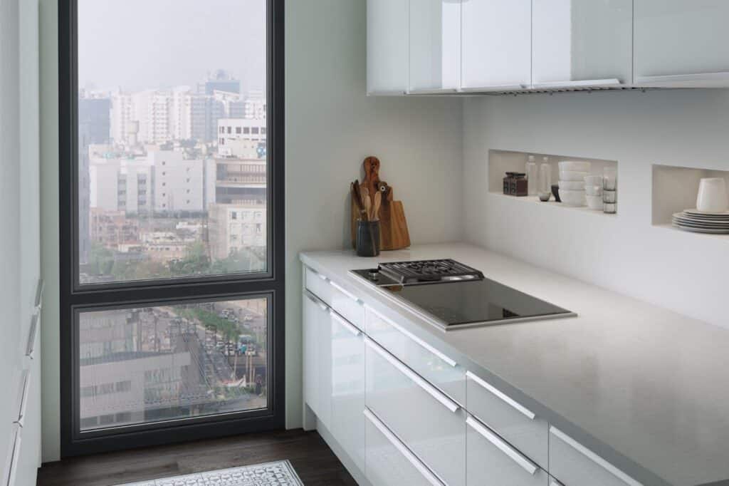 Modern white kitchen with a built-in induction range, utensils, and dishes on display shelves, featuring a large window overlooking an urban cityscape.