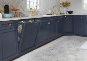 A modern kitchen with navy cabinets, gold hardware, a marble floor, and an open Cove Dishwasher seamlessly integrated into the cabinetry beneath the countertop.
