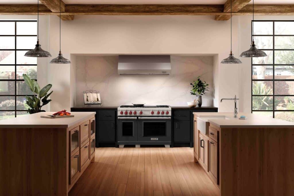 Modern kitchen with large gas range, highlighting the gas vs induction debate, two islands, pendant lights, wood floors, black cabinets, and large windows letting in natural light.