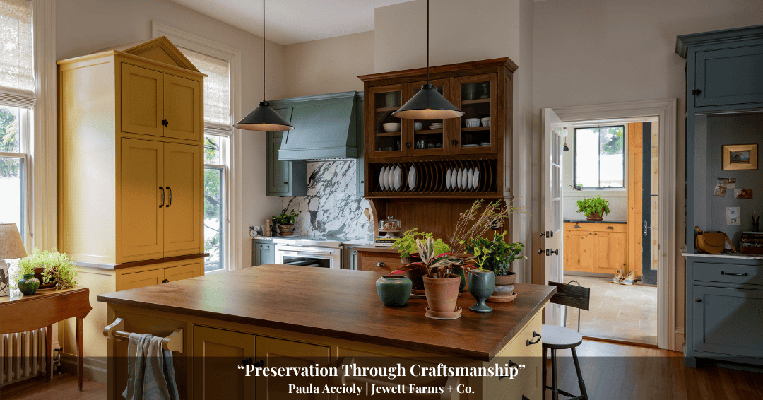 A bright, traditional kitchen with a wooden island, green and yellow cabinetry, open shelves, potted plants, and subtle touches of Clarke History, plus a door leading to another sunlit room.