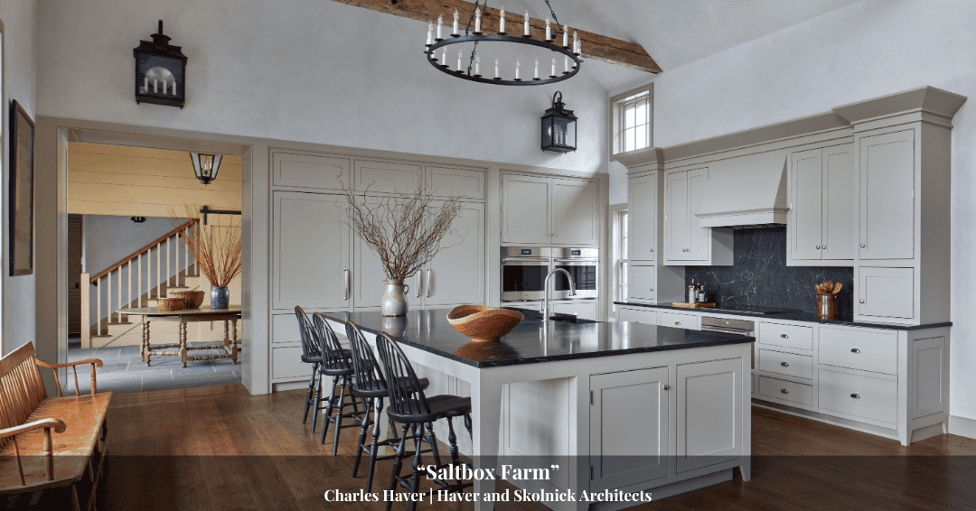 Spacious kitchen with white cabinetry, black island with stools, wooden beams, and pendant lights; seating and decorative vases visible in adjacent room, reflecting timeless style inspired by Clarke History.