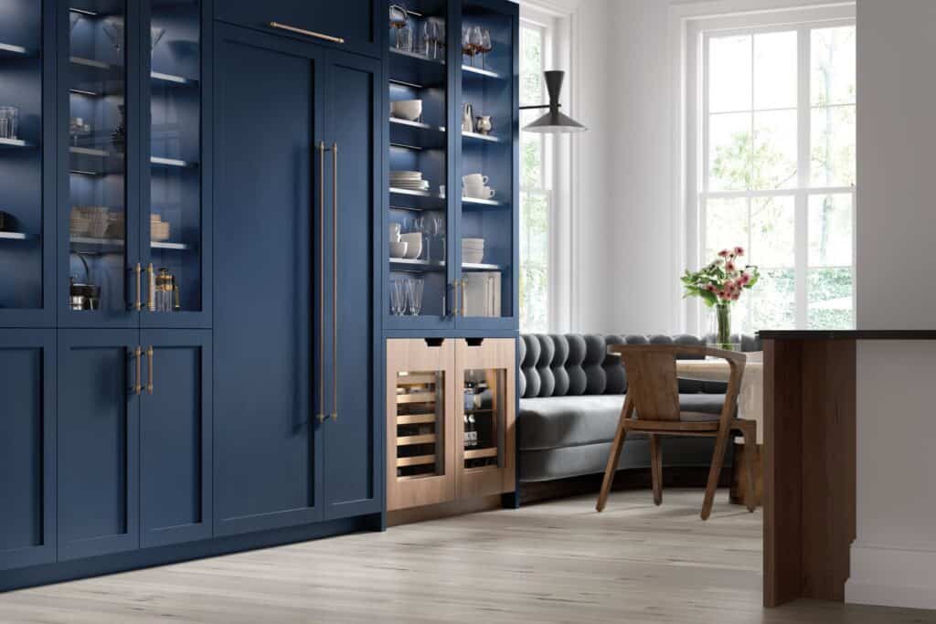 A modern kitchen with blue cabinets, glass doors, a built-in beverage refrigerator, and a dining nook featuring a wooden table and gray cushioned bench by large windows.