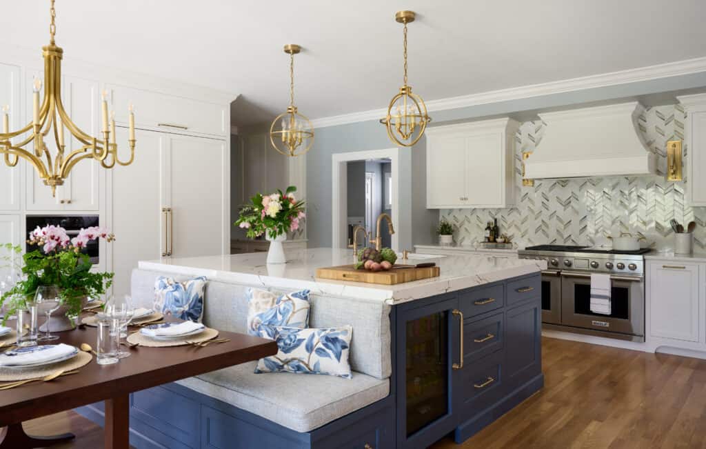 Modern kitchen from the Wenham Project features white cabinets, a blue island, built-in bench seating, gold light fixtures, wood floors, marble countertops, and stainless steel appliances.