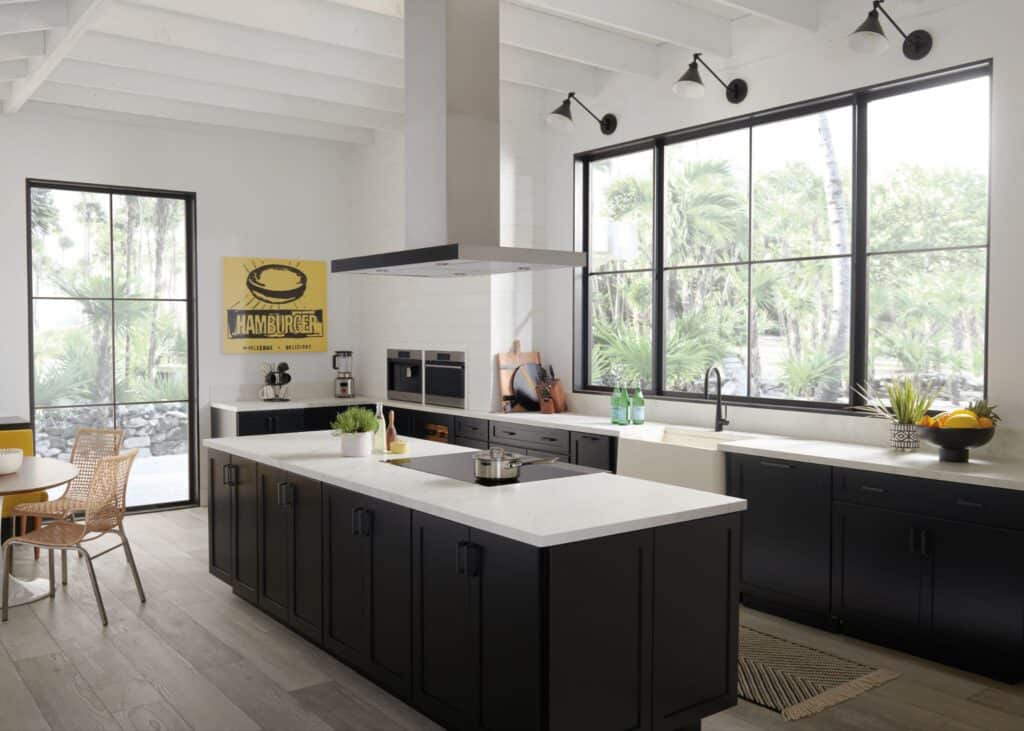Modern kitchen with black cabinets, white countertops, large windows, stainless steel appliances, and a central island featuring a sleek cooktop. A yellow poster and wicker chairs are visible in the background.