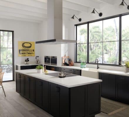 Modern kitchen with black cabinets, white countertops, large windows, stainless steel appliances, and a central island featuring a sleek cooktop. A yellow poster and wicker chairs are visible in the background.