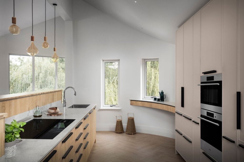 Modern kitchen with light wood cabinets, built-in appliances, a sleek cooktop, curved countertop, pendant lights, and large windows overlooking trees.