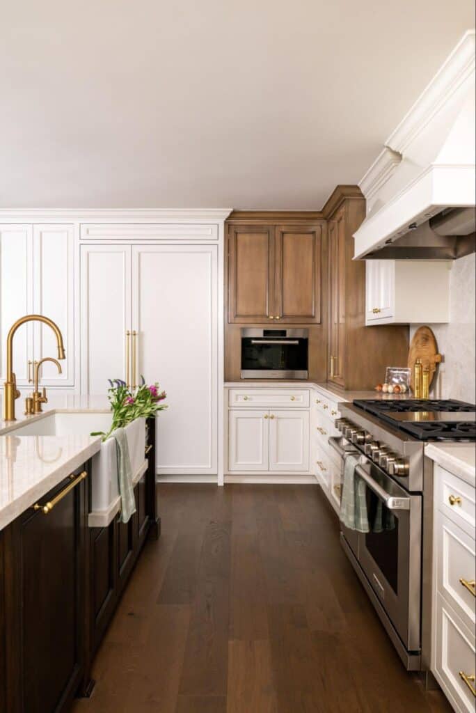 Modern kitchen with white and dark wood cabinetry, stainless steel appliances, gold fixtures, and a marble countertop with a farmhouse sink and fresh flowers.