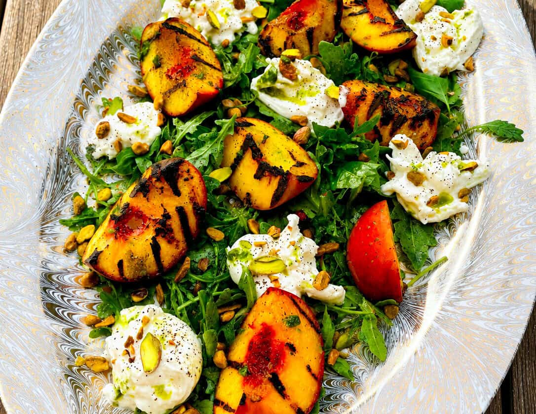 This vibrant grilled peach & burrata salad is beautifully arranged on an oval platter, featuring juicy grilled peaches, creamy burrata cheese, fresh arugula, crunchy pistachios, and fragrant herbs atop a rustic wooden table.