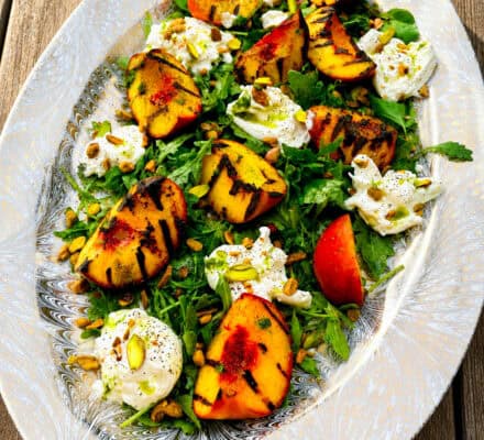 This vibrant grilled peach & burrata salad is beautifully arranged on an oval platter, featuring juicy grilled peaches, creamy burrata cheese, fresh arugula, crunchy pistachios, and fragrant herbs atop a rustic wooden table.