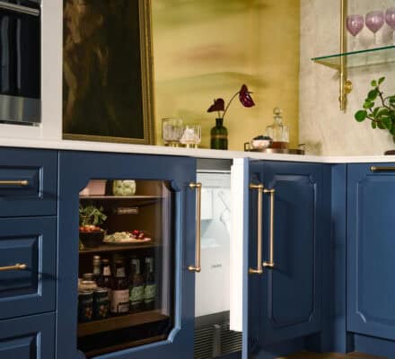 A kitchen with blue cabinets, a glass-front mini fridge stocked with drinks, an ice maker tucked neatly beside the counter, a partially open door, and decorative items on the counter and shelves.