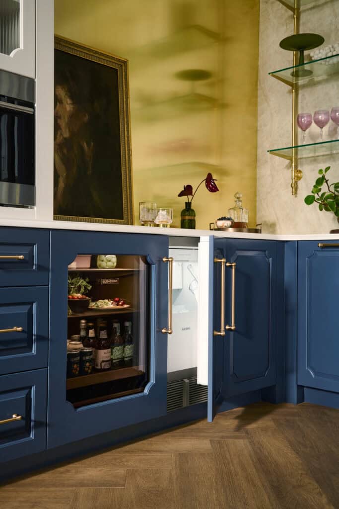 A kitchen with blue cabinets, a glass-front mini fridge stocked with drinks, an ice maker tucked neatly beside the counter, a partially open door, and decorative items on the counter and shelves.