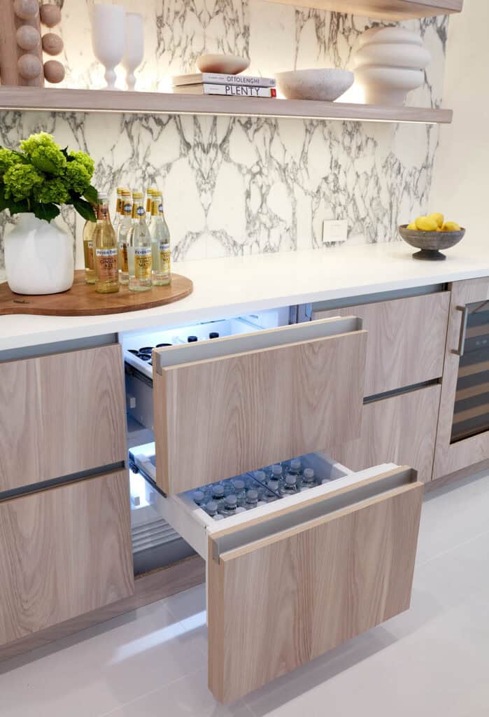 Two open refrigerator drawers in a modern kitchen, designed with the best kitchen layout for entertaining, reveal bottled drinks and water, while shelves above hold decor, a vase, and a bowl of lemons on the counter.