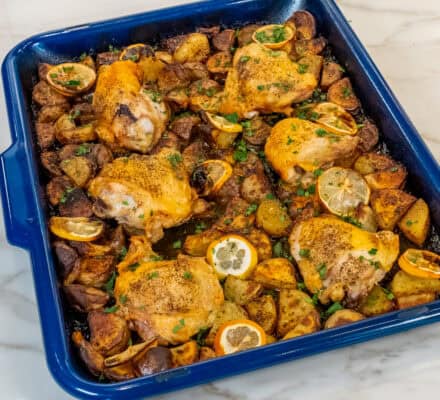 A blue baking dish with roasted chicken thighs, Greek lemon chicken flavors, sliced lemons, and seasoned potato chunks reminiscent of crispy lemon potatoes on a marble countertop.