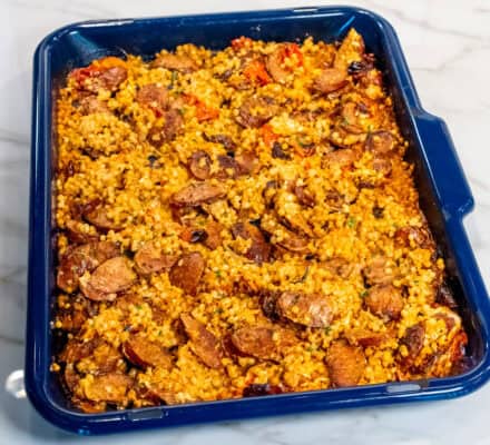 A blue baking dish filled with baked feta, sliced sausage, vegetables, and herbed couscous sits on a marble countertop.