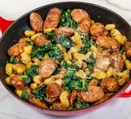 Red skillet containing crispy gnocchi, cooked sausage slices, and leafy greens, topped with grated cheese, on a marble countertop.