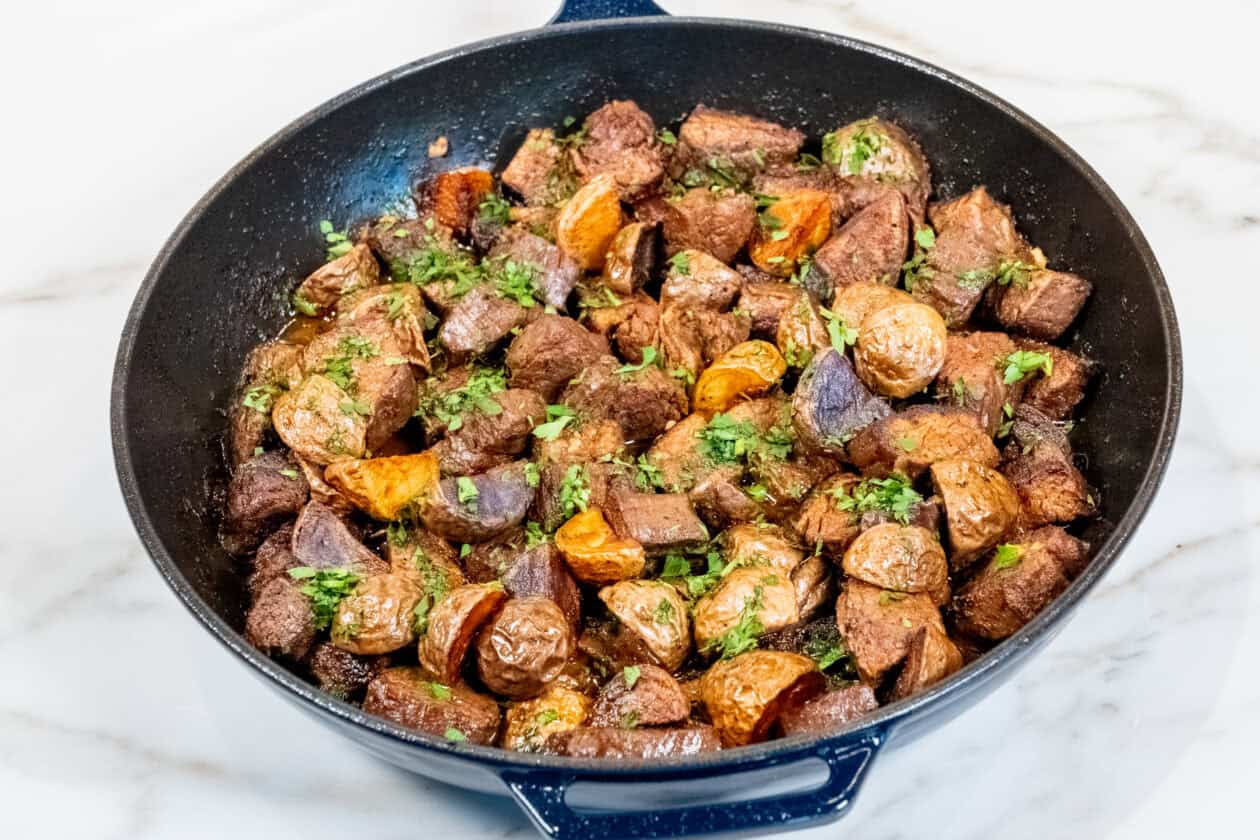 A skillet filled with garlic butter steak chunks and roasted potatoes, garnished with chopped herbs, on a marble countertop.