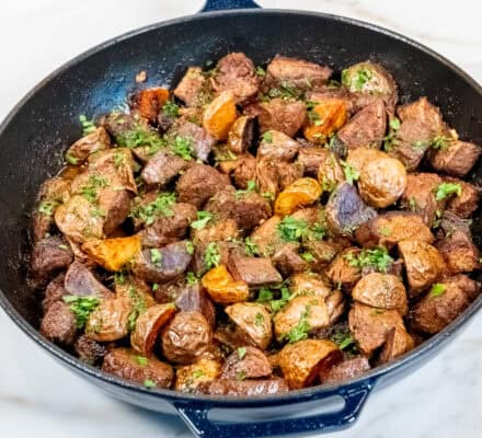 A skillet filled with garlic butter steak chunks and roasted potatoes, garnished with chopped herbs, on a marble countertop.