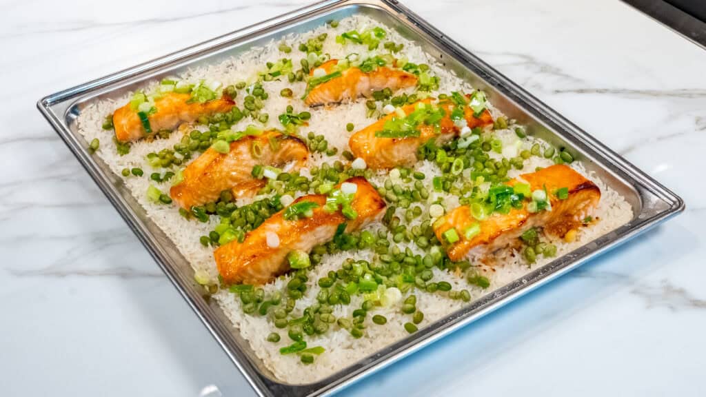 Six pieces of miso-glazed salmon rest on a baking sheet alongside fluffy coconut rice, tender snap peas, and chopped green onions on a marble countertop.