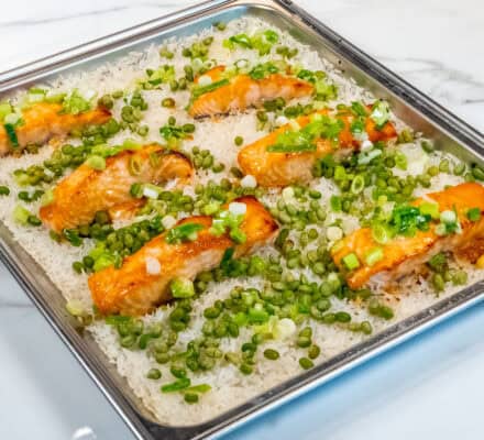 Six pieces of miso-glazed salmon rest on a baking sheet alongside fluffy coconut rice, tender snap peas, and chopped green onions on a marble countertop.