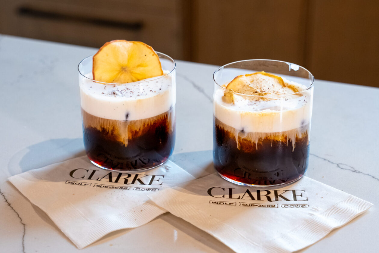 Two mont blanc iced coffees with cream and Vermont Apple Cider, garnished with apple slices on top, served in clear glasses on branded napkins on a marble countertop.