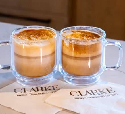 Two glass mugs of frothy honey tahini latte sit on a white countertop, each placed on a white napkin printed with the words “CLARKE WOLF SUBZERO COVE.”.