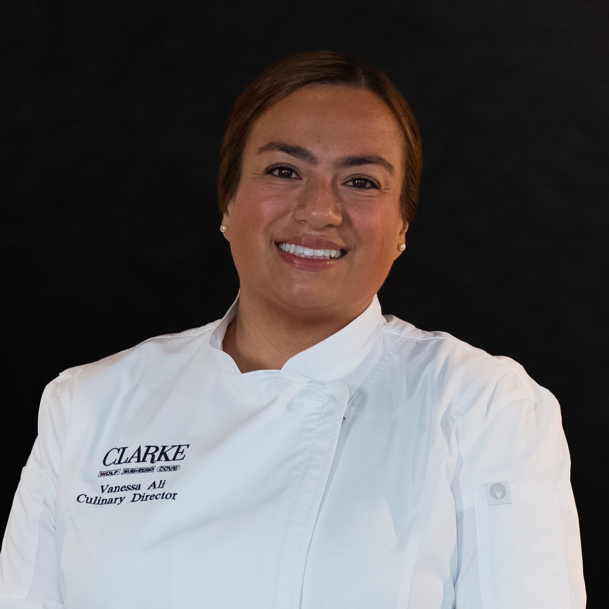 Meet the Clarke Showroom Team: A person wearing a white chef's coat with "Clarke" and "Culinary Director" embroidered on it stands against a plain dark background, smiling at the camera.